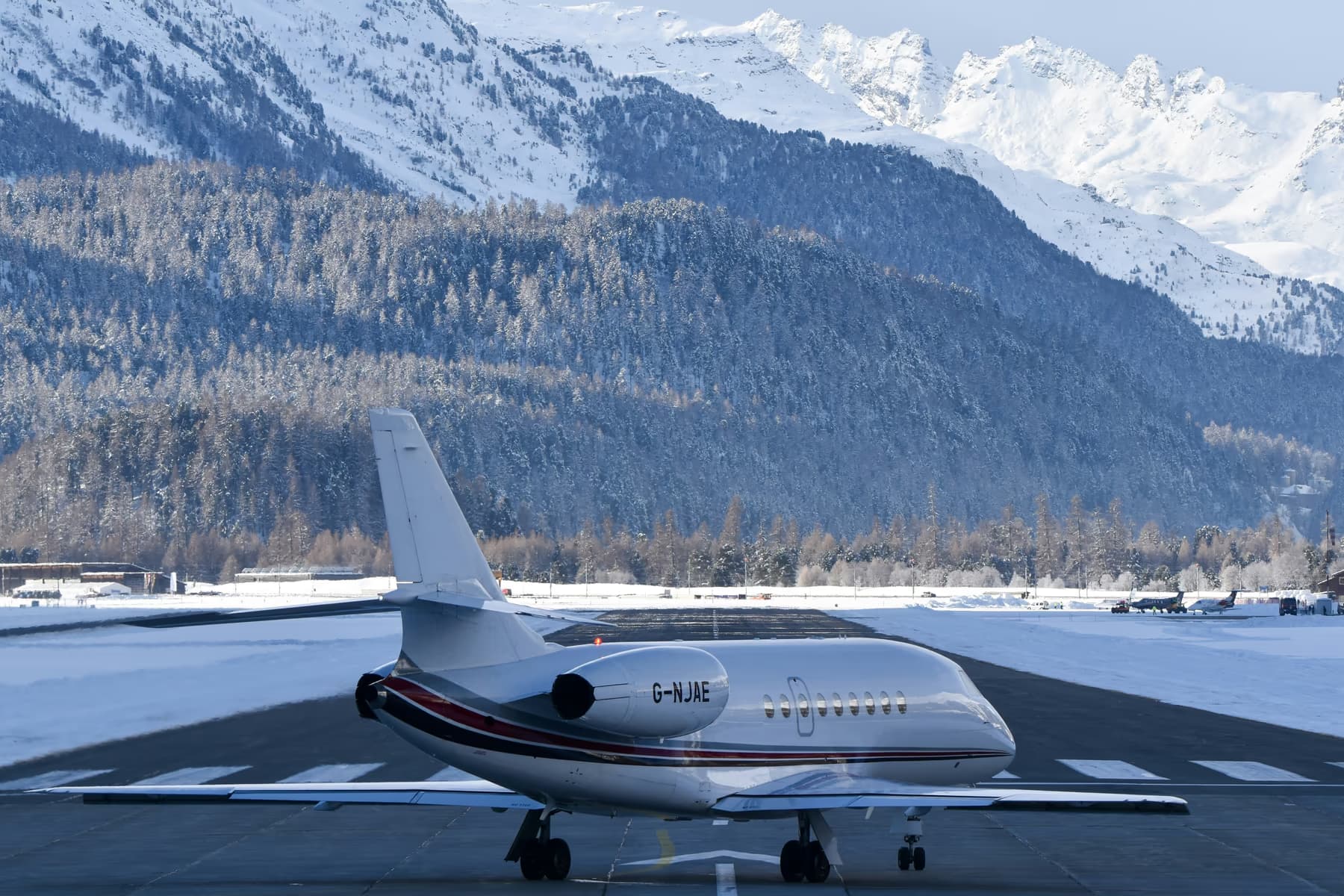 A Falcon private jet at rest on a plowed alpine apron, snow-capped mountains rising behind
